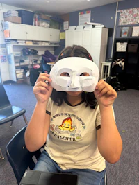 a girl holding a white mask in front of a laptop